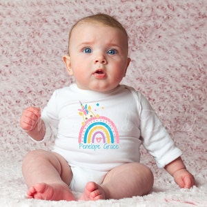 May include: A white baby onesie with a rainbow and unicorn design. The rainbow is multi-colored and has a heart in the middle. The text "Penelope Grace" is printed below the rainbow.
