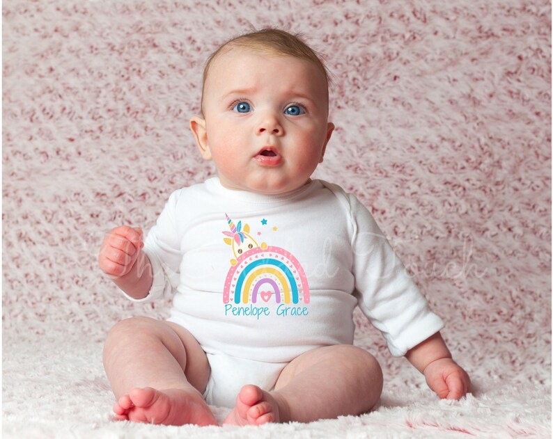May include: A white baby onesie with a rainbow and unicorn design. The rainbow is multi-colored and has a heart in the middle. The text "Penelope Grace" is printed below the rainbow.