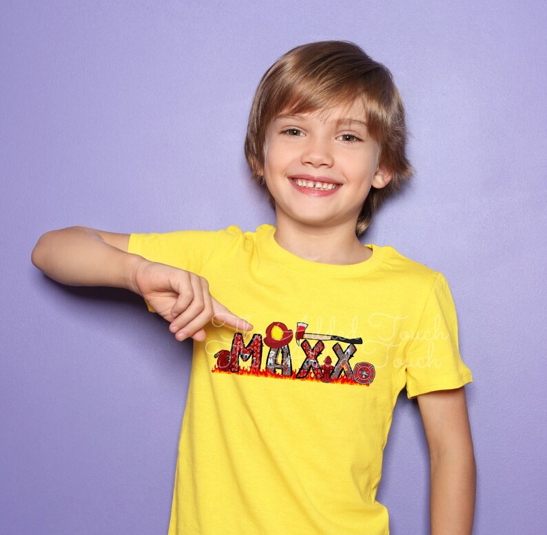 May include: A young boy wearing a yellow t-shirt with a firefighter themed design that says "MAXX" in red, black, and white. The design includes a fire helmet, axe, and hose.