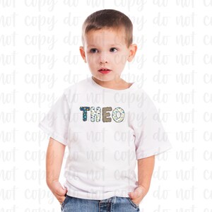 K&ouml;nnte beinhalten: Ein wei&szlig;es T-Shirt mit einem blauen, gr&uuml;nen und braunen gemusterten Namen "THEO" auf der Vorderseite. Ein kleiner Junge tr&auml;gt das Shirt und hat die H&auml;nde in den Taschen.