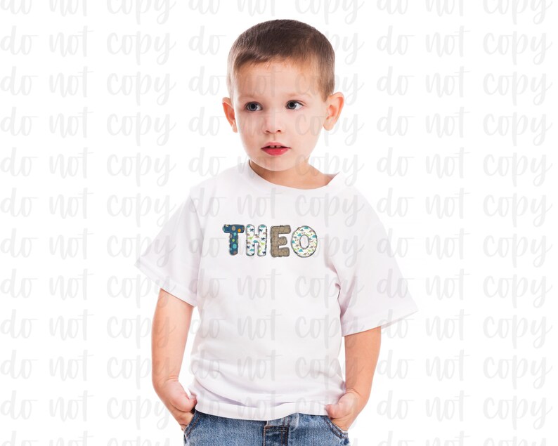 K&ouml;nnte beinhalten: Ein wei&szlig;es T-Shirt mit einem blauen, gr&uuml;nen und braunen gemusterten Namen "THEO" auf der Vorderseite. Ein kleiner Junge tr&auml;gt das Shirt und hat die H&auml;nde in den Taschen.