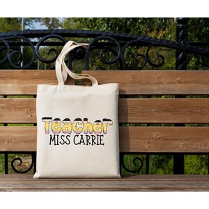 May include: A white canvas tote bag with black text that reads "Teacher Miss Carrie". The text is stylized to look like a pencil. The bag is sitting on a wooden bench.