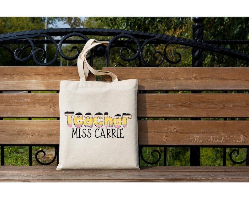 May include: A white canvas tote bag with black text that reads "Teacher Miss Carrie". The text is stylized to look like a pencil. The bag is sitting on a wooden bench.