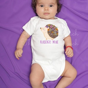May include: White baby onesie with a hedgehog graphic wearing a floral crown. The text "KALEIGH MAE" is printed below the hedgehog. The onesie is on a purple background.