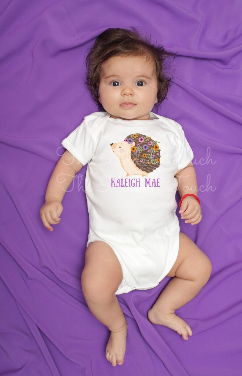 May include: White baby onesie with a hedgehog graphic wearing a floral crown. The text "KALEIGH MAE" is printed below the hedgehog. The onesie is on a purple background.