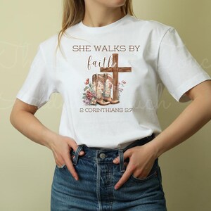 May include: White t-shirt with the text "She walks by faith" and a pair of brown cowboy boots with a cross behind them. The text "2 Corinthians 5:7" is below the boots.