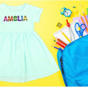 May include: A light blue dress with the name "Amelia" spelled out in colorful letters. The dress is next to a blue backpack filled with school supplies, including pencils, a ruler, a compass, and a pair of scissors.