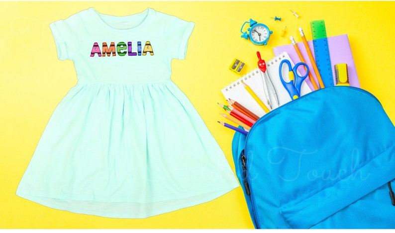 May include: A light blue dress with the name "Amelia" spelled out in colorful letters. The dress is next to a blue backpack filled with school supplies, including pencils, a ruler, a compass, and a pair of scissors.