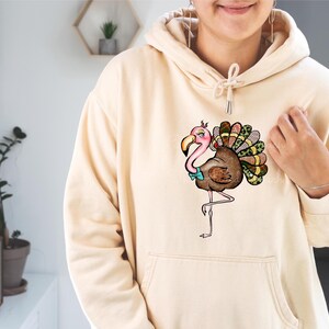 May include: A beige hoodie featuring a whimsical illustration of a turkey with flamingo-like features. The turkey has a pink head and beak, a brown body, and a colorful tail. It is wearing a blue bow tie. The hoodie has a front pocket and a drawstring hood.