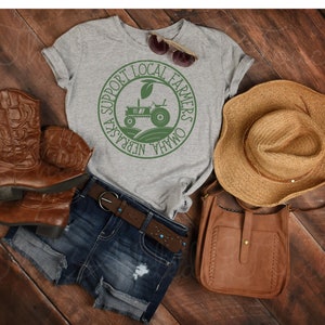 Pu&ograve; includere: Una maglietta grigia con una grafica circolare verde che dice "Support Local Farmers Omaha Nebraska" con un trattore all'interno del cerchio. La maglietta &egrave; appoggiata su una superficie di legno con un paio di stivali da cowboy marroni, una borsa di pelle marrone e un cappello di paglia.