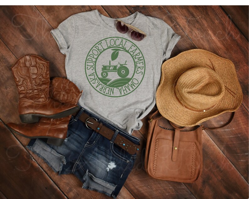 Pu&ograve; includere: Una maglietta grigia con una grafica circolare verde che dice "Support Local Farmers Omaha Nebraska" con un trattore all'interno del cerchio. La maglietta &egrave; appoggiata su una superficie di legno con un paio di stivali da cowboy marroni, una borsa di pelle marrone e un cappello di paglia.