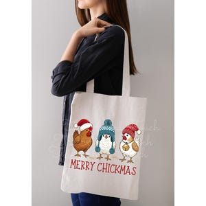 May include: White tote bag featuring three cartoon chickens wearing winter hats. The chickens are brown, white, and blue. The text "MERRY CHICKMAS" is printed in red below the chickens. The bag is held by a person.