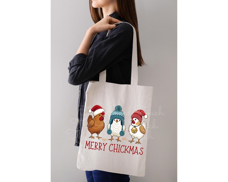 May include: White tote bag featuring three cartoon chickens wearing winter hats. The chickens are brown, white, and blue. The text "MERRY CHICKMAS" is printed in red below the chickens. The bag is held by a person.