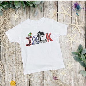 May include: White short-sleeved t-shirt with the name "JACK" spelled out in colorful letters with a pirate theme. The letters are decorated with a parrot, a pirate hat, and stripes.
