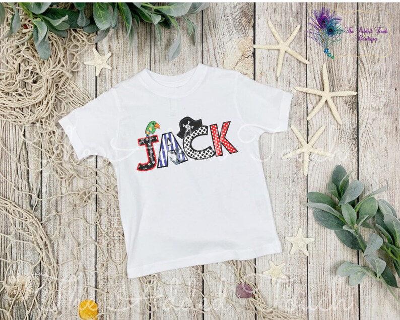 May include: White short-sleeved t-shirt with the name "JACK" spelled out in colorful letters with a pirate theme. The letters are decorated with a parrot, a pirate hat, and stripes.