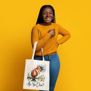 Pode incluir: Uma mulher est&aacute; segurando uma bolsa tote branca com uma ilustra&ccedil;&atilde;o de um p&aacute;ssaro de desenho animado e o texto "Are You Okay?" O p&aacute;ssaro &eacute; vermelho, laranja e marrom com um bico azul. A bolsa tem uma al&ccedil;a branca.