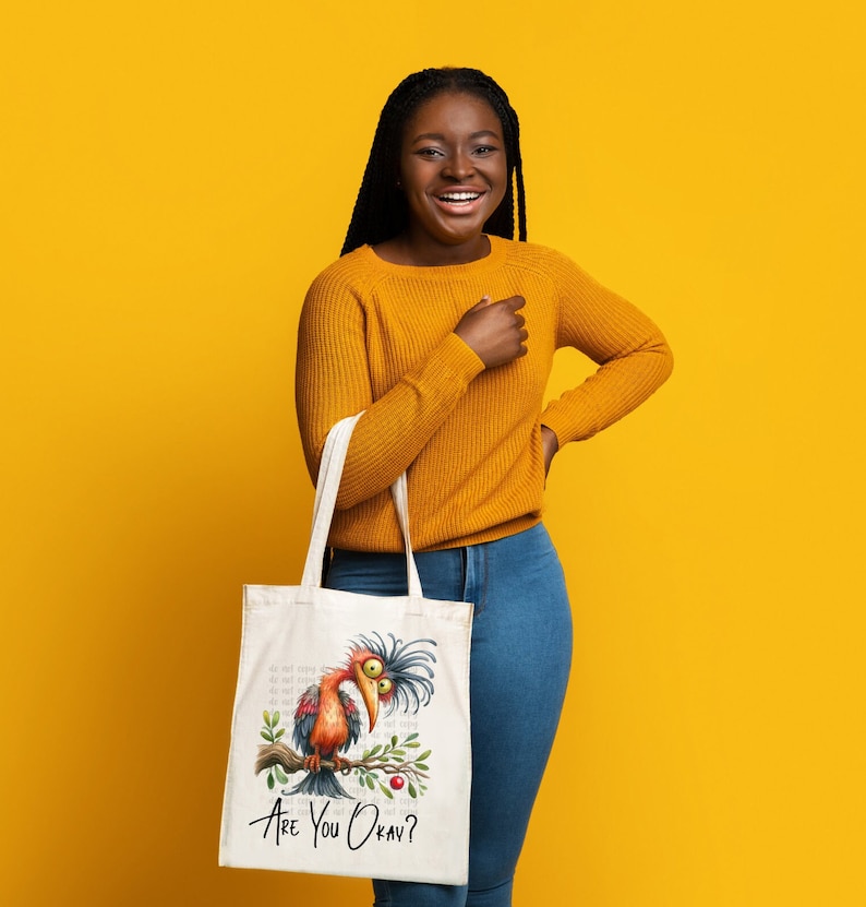 Pode incluir: Uma mulher est&aacute; segurando uma bolsa tote branca com uma ilustra&ccedil;&atilde;o de um p&aacute;ssaro de desenho animado e o texto "Are You Okay?" O p&aacute;ssaro &eacute; vermelho, laranja e marrom com um bico azul. A bolsa tem uma al&ccedil;a branca.