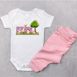 May include: A white baby onesie with a pink and white polka dot design featuring the name "FERN" in pink letters. The onesie has a farm animal theme with a chick, pig, and cow. A pink tree with green leaves is in the background. The onesie is paired with pink pants.