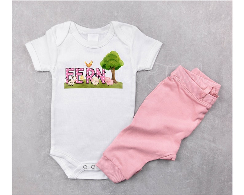 May include: A white baby onesie with a pink and white polka dot design featuring the name "FERN" in pink letters. The onesie has a farm animal theme with a chick, pig, and cow. A pink tree with green leaves is in the background. The onesie is paired with pink pants.