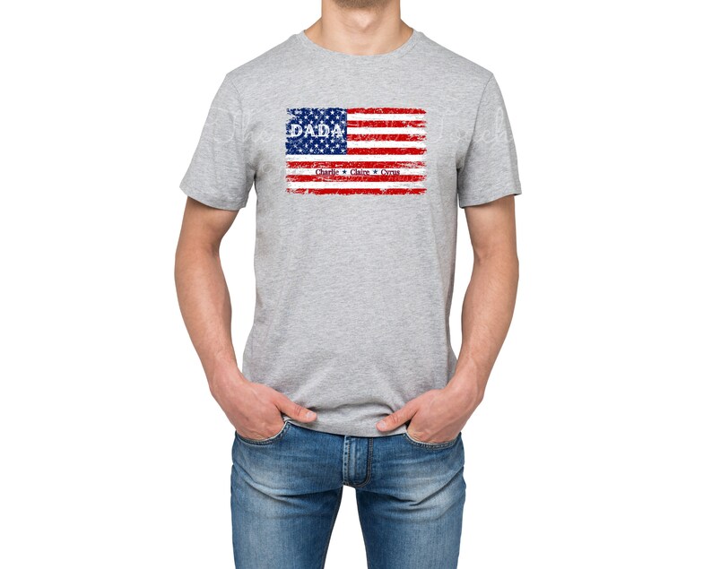 May include: A gray t-shirt with a distressed American flag design. The flag has the word "DADA" in blue letters and the words "Charlie, Claire & Cyrus" in smaller blue letters below the flag.