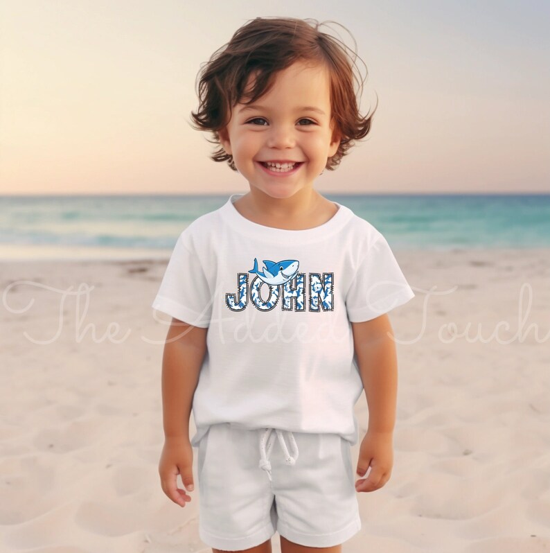 May include: A white t-shirt with a blue shark graphic and the name "JOHN" spelled out in blue and white letters.