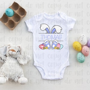 May include: White baby onesie with a blue bunny rabbit peeking over the top. The bunny has purple ears and blue feet. The onesie has the name "THOMAS" printed on it. There are colorful Easter eggs around the bunny.