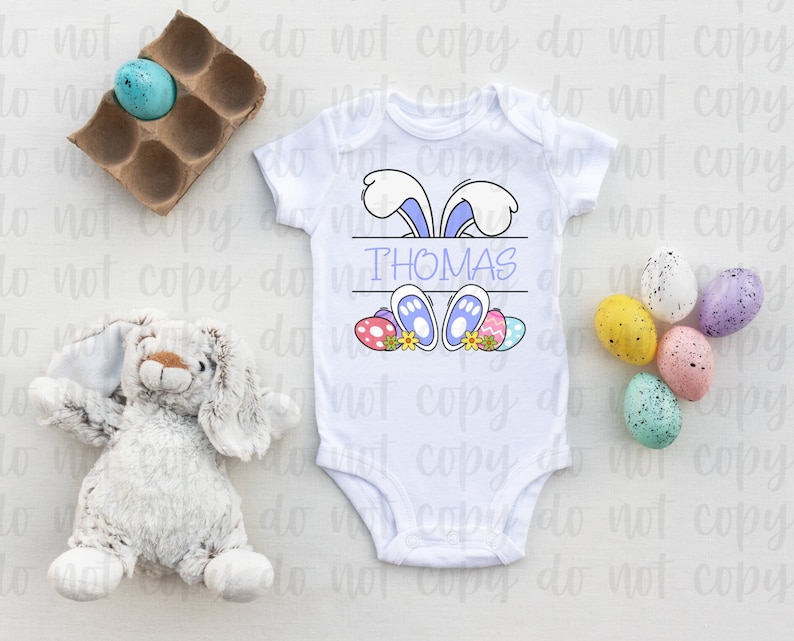 May include: White baby onesie with a blue bunny rabbit peeking over the top. The bunny has purple ears and blue feet. The onesie has the name "THOMAS" printed on it. There are colorful Easter eggs around the bunny.