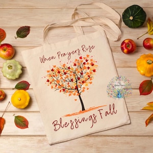 May include: A cream tote bag featuring a tree design and the words "When Prayers Rise Blessings Fall." The bag is surrounded by pumpkins, gourds, apples, and autumn leaves on a wooden surface. The image has a harvest theme.