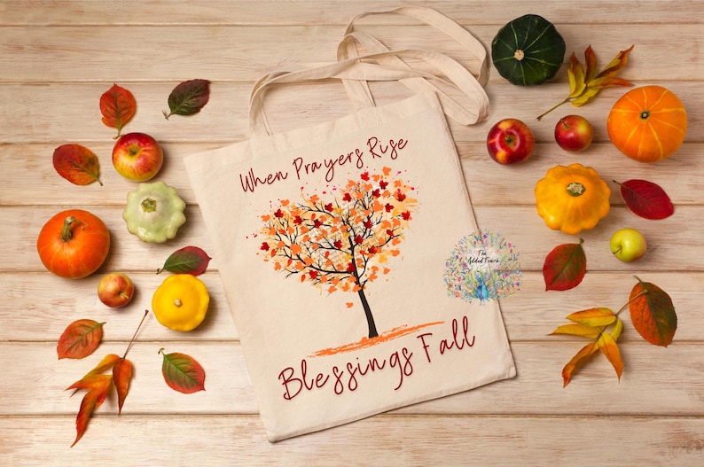 May include: A cream tote bag featuring a tree design and the words "When Prayers Rise Blessings Fall." The bag is surrounded by pumpkins, gourds, apples, and autumn leaves on a wooden surface. The image has a harvest theme.