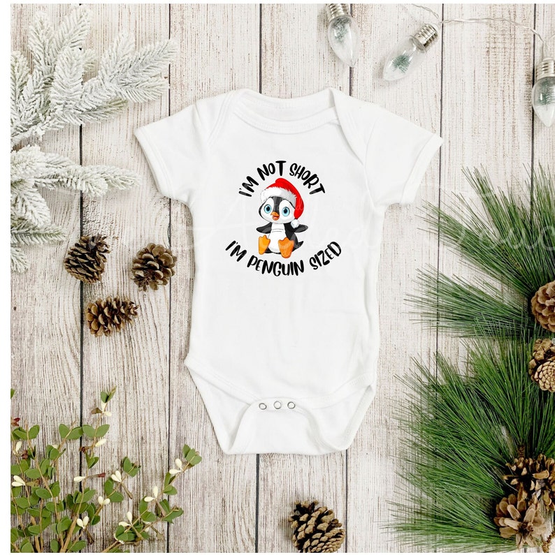 May include: White baby onesie with a graphic of a penguin wearing a Santa hat. The text on the onesie reads "I'm not short, I'm penguin sized." The onesie is surrounded by pinecones, greenery, and decorative lights.