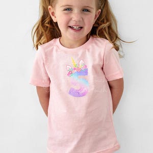 May include: Pink short-sleeved t-shirt featuring a unicorn design with a floral mane and a golden horn. The number 5 is integrated into the design, with the name "Tracy" below. The shirt is worn by a young girl.