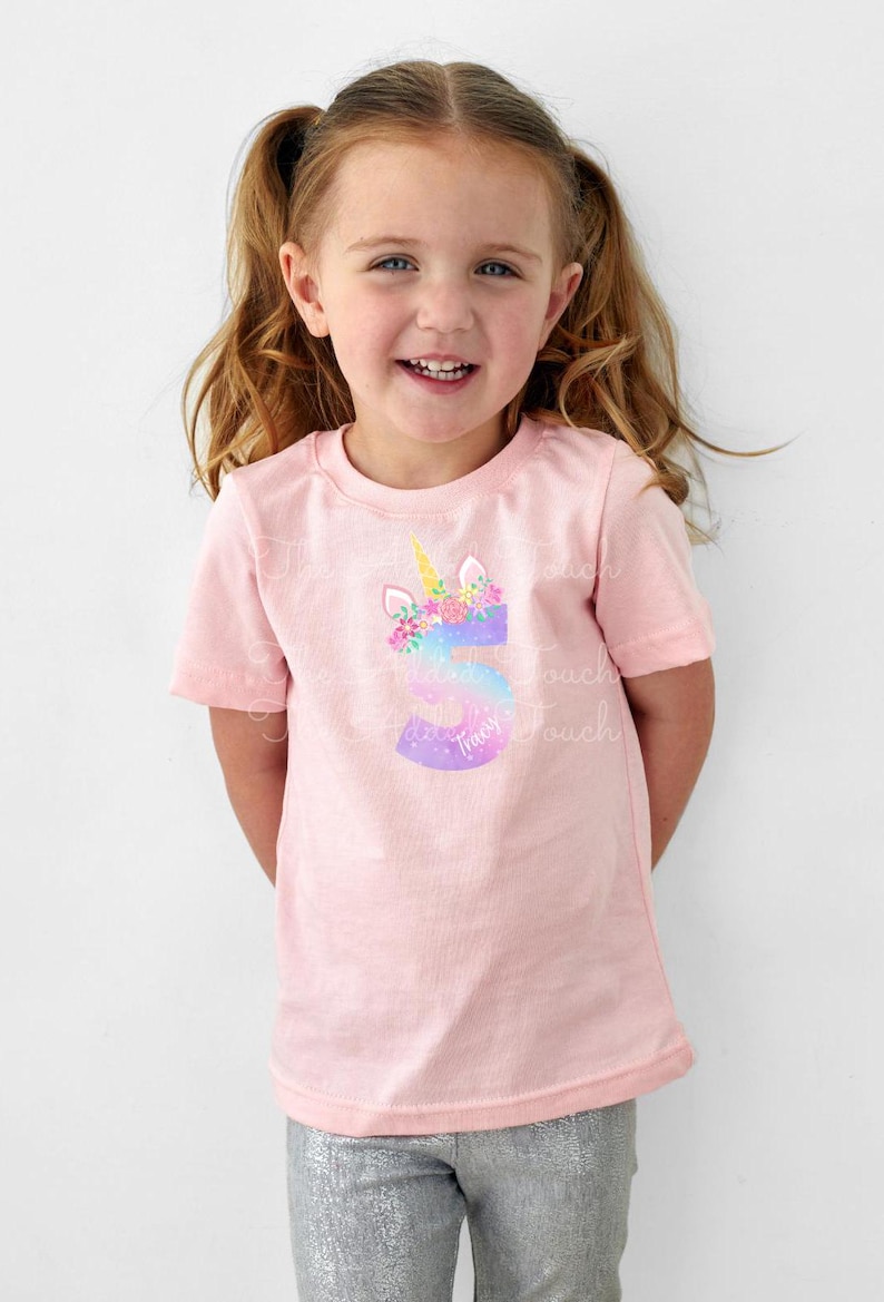 May include: Pink short-sleeved t-shirt featuring a unicorn design with a floral mane and a golden horn. The number 5 is integrated into the design, with the name "Tracy" below. The shirt is worn by a young girl.