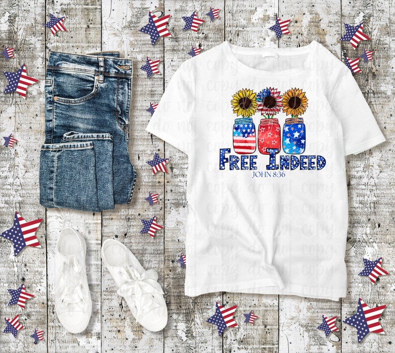 May include: White t-shirt featuring a design of three mason jars decorated with red, white and blue stars and sunflowers. The text "Free Indeed" and "John 8:36" is printed below the jars.