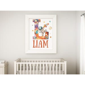 May include: A white framed print with a watercolor illustration of a letter L with an astronaut in a space suit holding a rocket. The letter L is filled with a galaxy design and the name Liam is printed below the letter.