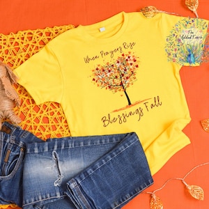 May include: Yellow t-shirt with the text "When Prayers Rise Blessings Fall" and a tree design. The image also includes blue jeans and a small doll. The background is orange.