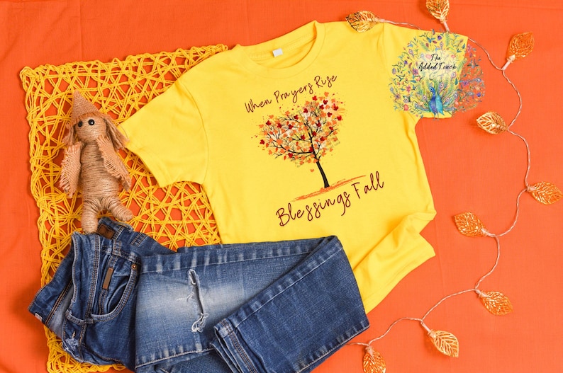 May include: Yellow t-shirt with the text "When Prayers Rise Blessings Fall" and a tree design. The image also includes blue jeans and a small doll. The background is orange.