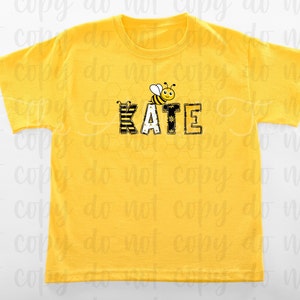 May include: Yellow short-sleeved t-shirt with a black and yellow bee graphic and the name "KATE" in black letters with a yellow and black striped pattern.