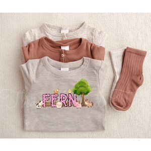 May include: A light brown, grey, and brown baby onesie with a pink and white polka dot design that says "FERN" with farm animals and a green tree. The onesie is folded and laid on a beige surface with two brown socks.