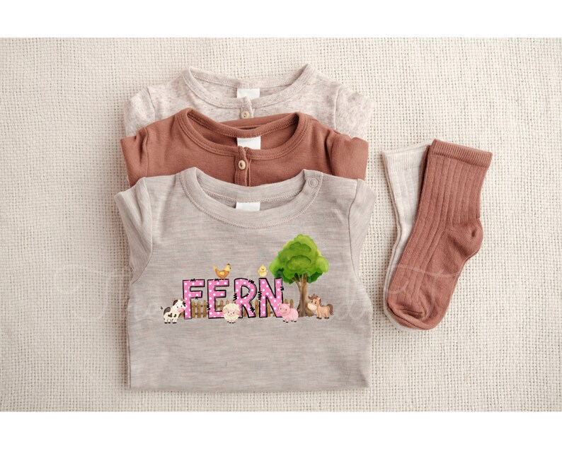 May include: A light brown, grey, and brown baby onesie with a pink and white polka dot design that says "FERN" with farm animals and a green tree. The onesie is folded and laid on a beige surface with two brown socks.