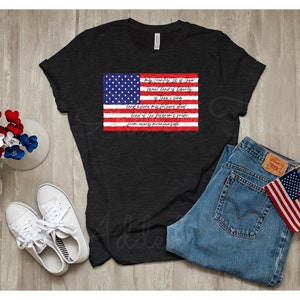 May include: A black t-shirt with a distressed American flag graphic and the lyrics to "My Country 'Tis of Thee" printed on it. The shirt is laid out on a wooden surface with a pair of white sneakers, a pair of blue jeans, and a small American flag.