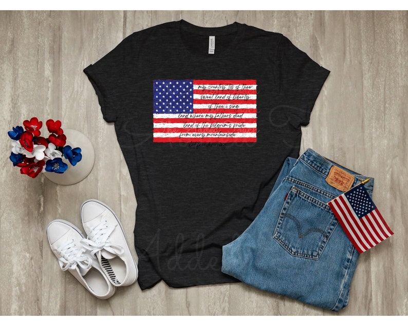 May include: A black t-shirt with a distressed American flag graphic and the lyrics to "My Country 'Tis of Thee" printed on it. The shirt is laid out on a wooden surface with a pair of white sneakers, a pair of blue jeans, and a small American flag.