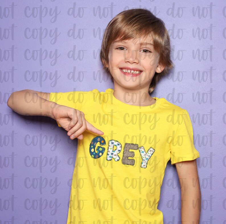 K&ouml;nnte beinhalten: Ein kleiner Junge tr&auml;gt ein gelbes T-Shirt mit dem Namen GREY, der in bunten Stoffbuchstaben geschrieben ist. Die Buchstaben sind mit einem Weltraum-Motiv verziert.
