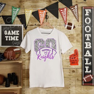 May include: White t-shirt with a purple number 60 with a leopard print design and a football with a heart. The text "Knights" is below the football.