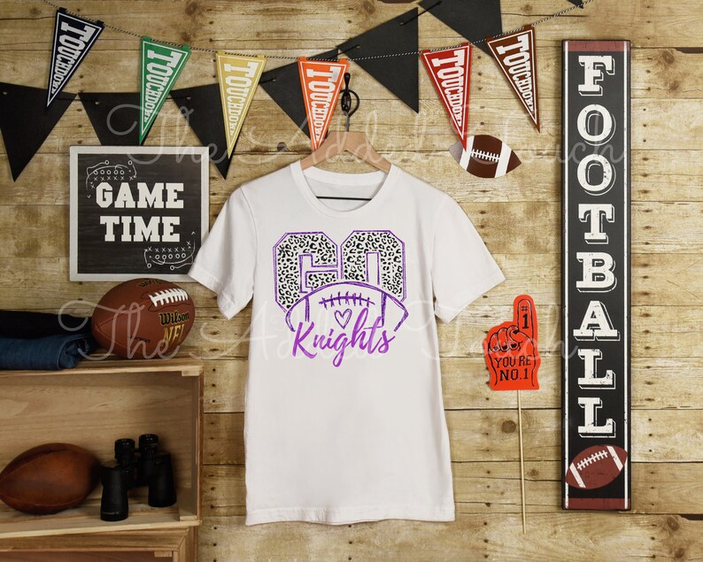 May include: White t-shirt with a purple number 60 with a leopard print design and a football with a heart. The text "Knights" is below the football.
