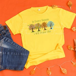 May include: A yellow t-shirt with a design of four trees in different shades of green, yellow, and orange. The trees are surrounded by leaves and the text "In Every Season I Will Praise Him".