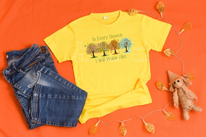 May include: A yellow t-shirt with a design of four trees in different shades of green, yellow, and orange. The trees are surrounded by leaves and the text "In Every Season I Will Praise Him".