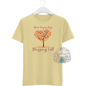 May include: A pale yellow t-shirt featuring the text "When Prayers Rise Blessings Fall" and a tree design with a heart-shaped crown of orange and red leaves. A floral design is on the lower right.