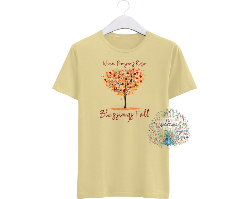 May include: A pale yellow t-shirt featuring the text "When Prayers Rise Blessings Fall" and a tree design with a heart-shaped crown of orange and red leaves. A floral design is on the lower right.