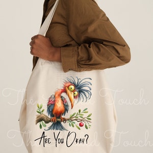 Pode incluir: Uma bolsa tote de lona branca com uma ilustra&ccedil;&atilde;o de um p&aacute;ssaro de desenho animado e o texto "Are you okay?" impresso nela. O p&aacute;ssaro tem penas azuis e laranja e est&aacute; empoleirado em um galho com uma baga vermelha.