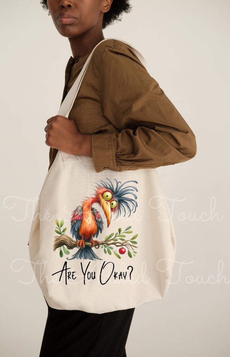 Pode incluir: Uma bolsa tote de lona branca com uma ilustra&ccedil;&atilde;o de um p&aacute;ssaro de desenho animado e o texto "Are you okay?" impresso nela. O p&aacute;ssaro tem penas azuis e laranja e est&aacute; empoleirado em um galho com uma baga vermelha.